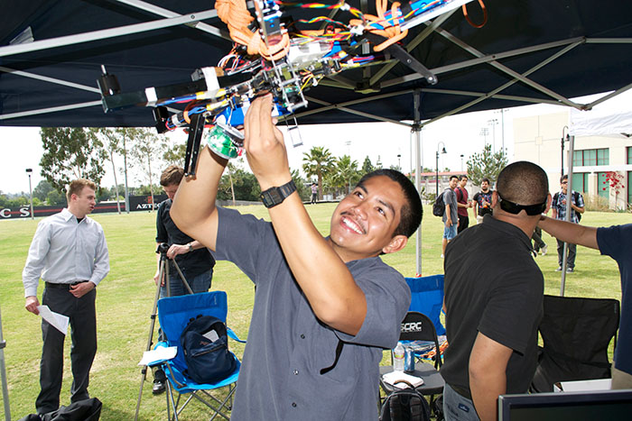 An engineering student is under a tent outdoors for Senior Design Day, holding up a complex, multi-colored drone with visible wires and components. The drone appears to be in the process of demonstration or testing. 