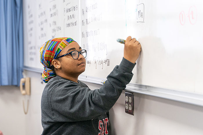 A young woman stands in front of a whiteboard, writing physics equations with a marker.