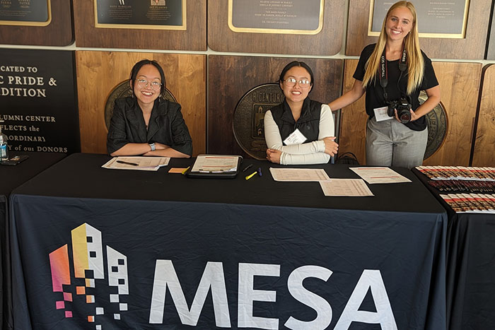 Three MESA staff members sit at a reception table waiting to offer assistance