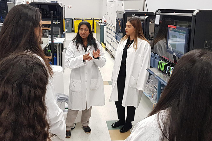 A group of MESA students in a laboratory setting, wearing white lab coats, listening to an industry professional. The background features various pieces of technical equipment and monitors on racks, suggesting an environment of scientific research or testing.